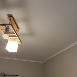 Molart - Szare wnętrze pokoju z drewnianą lampą sufitową i listwą dekoracyjną pod sufitem. Widoczny fragment okna z zasłoną.