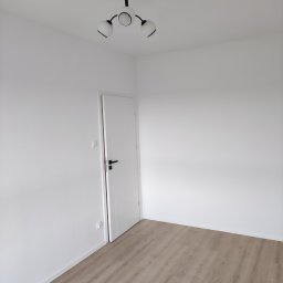 Molart - Jasne panele podłogowe w minimalistycznym pokoju z białymi ścianami, czarną lampą sufitową i białymi drzwiami z czarną klamką. Nowoczesne wykończenie.