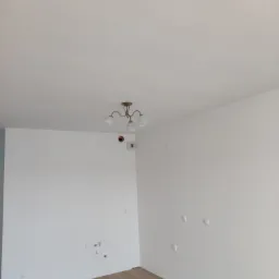 Jasne, odnowione wnętrze pokoju z panelami podłogowymi i białymi ścianami. Widoczna instalacja elektryczna i oświetlenie sufitowe.