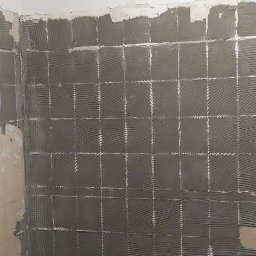 Molart - Ściana w trakcie układania płytek, widoczne klejone kwadraty i stos płytek na podłodze. Fragment ściany przygotowany pod płytki, sufit biały.