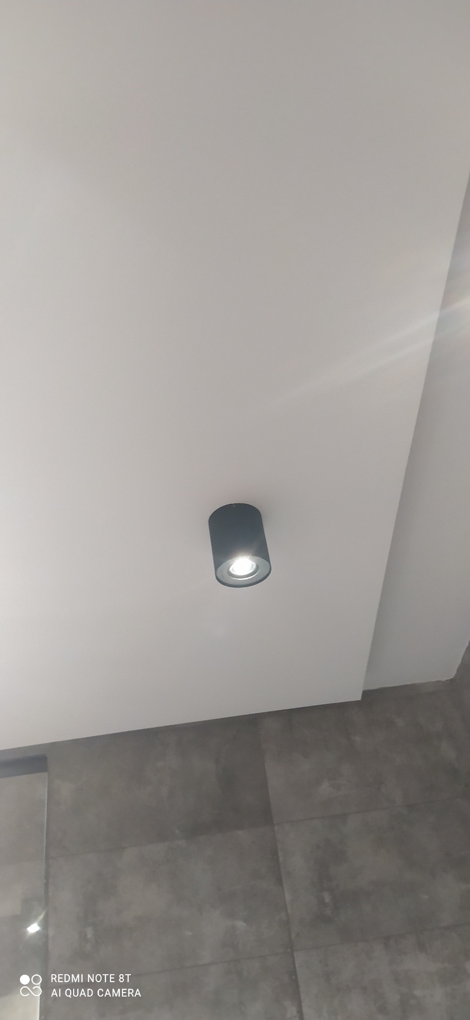 Czarna lampa sufitowa typu spot, zamontowana w białym suficie podwieszanym, widoczna na tle szarych płytek ściennych.