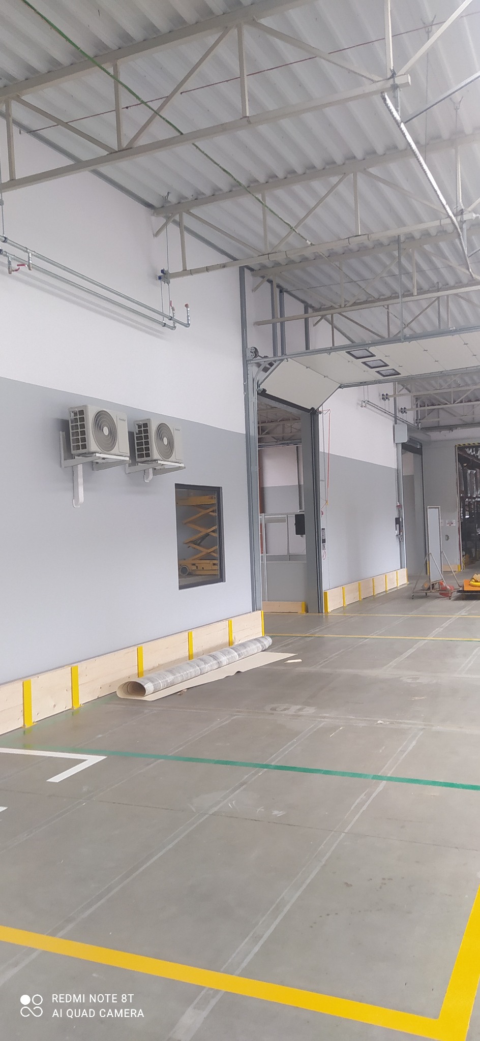 Wnętrze hali przemysłowej z szarymi ścianami, dwoma klimatyzatorami, żółto-zielonymi liniami na podłodze, drewnianą listwą przypodłogową i zwiniętym dywanem przy ścianie.