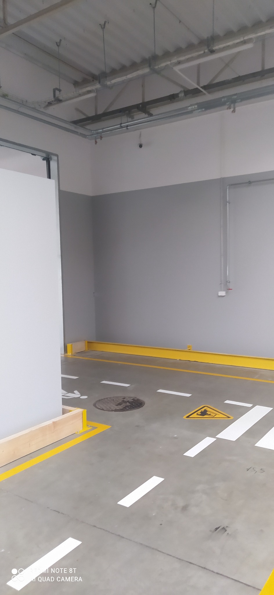 Oznakowanie poziome w hali: żółte bariery, białe pasy przejścia dla pieszych, symbol ostrzegawczy i piktogram osoby niepełnosprawnej na szarym, betonowym podłożu.