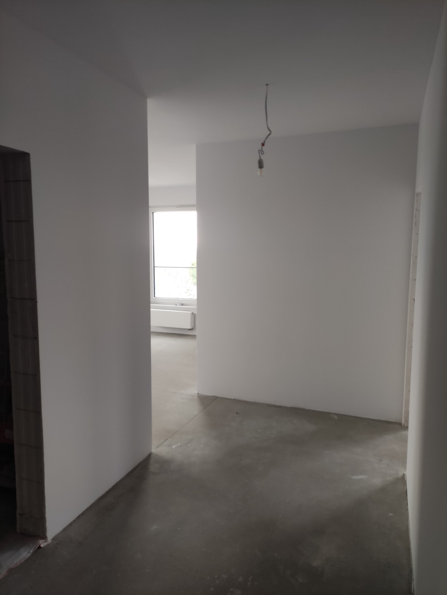 Wnętrze po szpachlowaniu i malowaniu na biało. Widoczna surowa podłoga i wisząca żarówka. W głębi okno z grzejnikiem. Stan wykończeniowy.