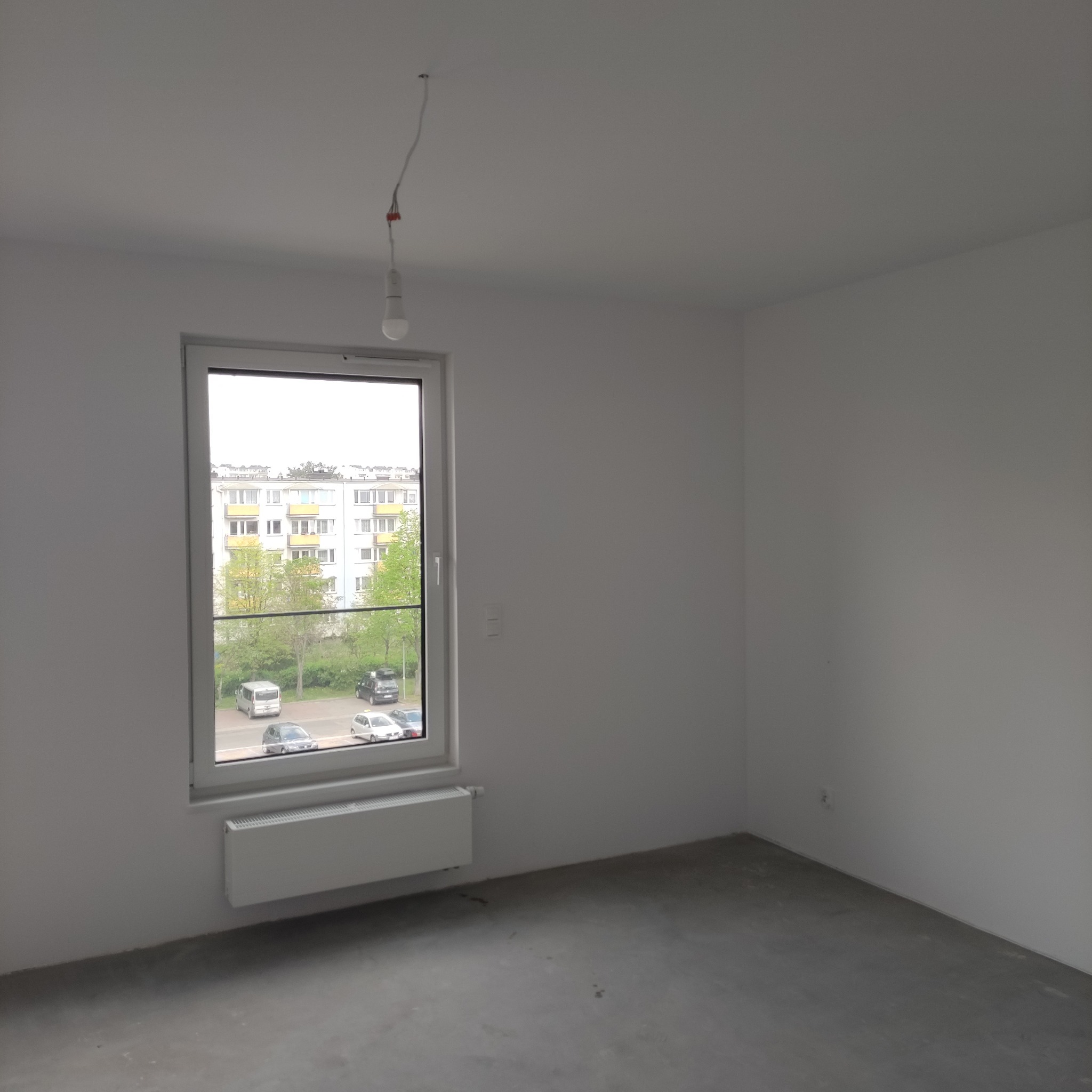 Wnętrze pokoju w stanie deweloperskim z oknem, grzejnikiem i surową żarówką. Ściany białe, podłoga szara. Widok na blok mieszkalny za oknem.