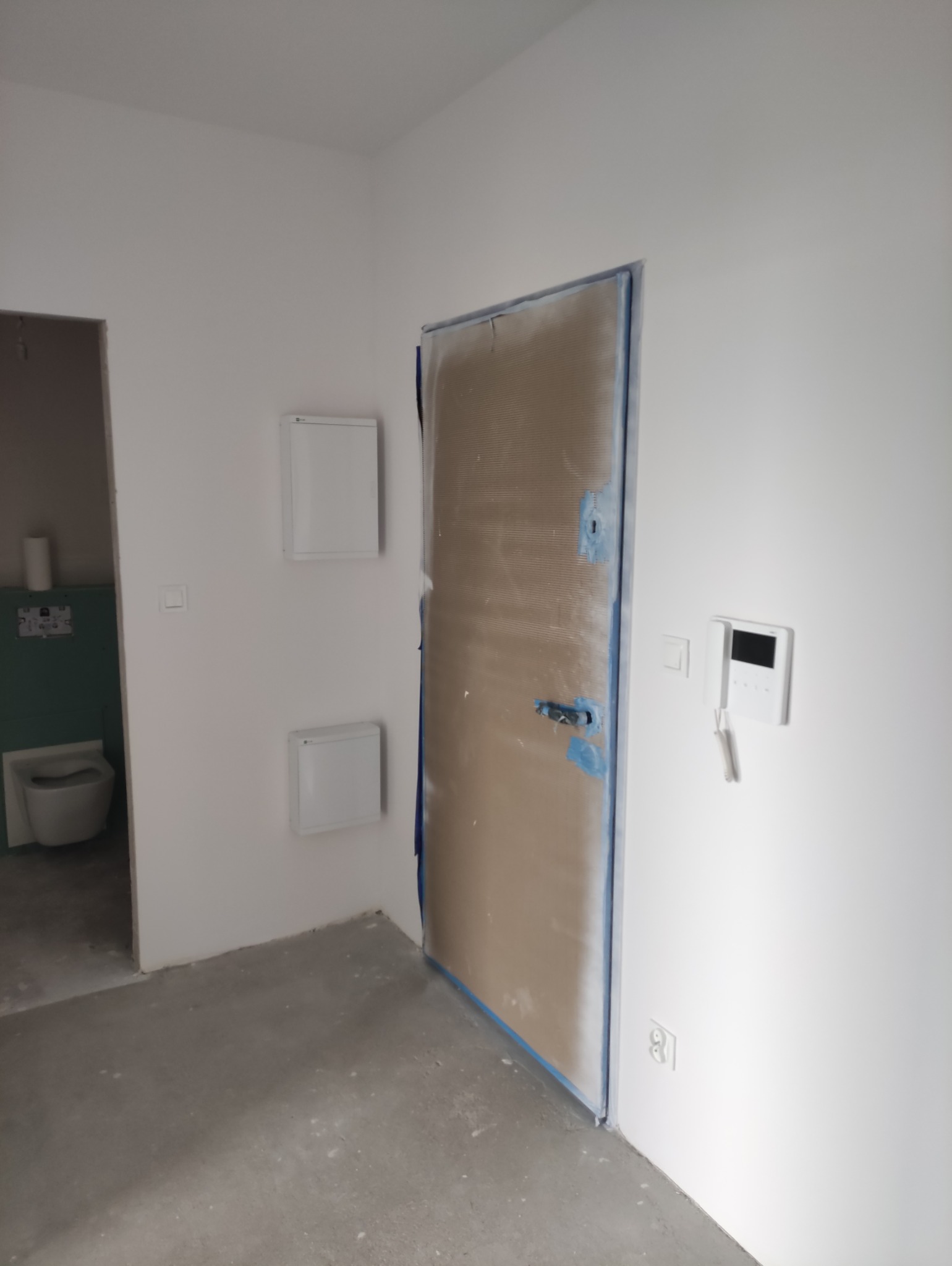 Wnętrze w trakcie remontu: zabezpieczone drzwi folią malarską, ściany po szpachlowaniu, widoczne puszki instalacyjne i fragment łazienki w tle. Surowy stan wykończenia.