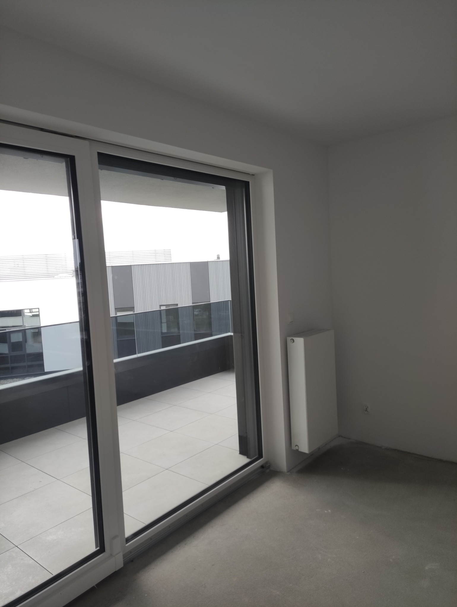 Jasne wnętrze z widokiem na balkon przez przesuwne drzwi. Ściany w kolorze złamanej bieli, widoczny grzejnik. Na zewnątrz nowoczesny budynek.