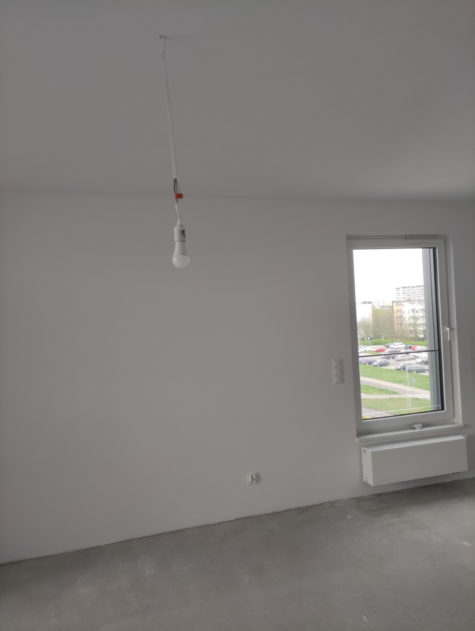 Surowe wnętrze pokoju z szarą wylewką, białymi ścianami i oknem z widokiem na parking. Tymczasowa żarówka zwisa z sufitu.
