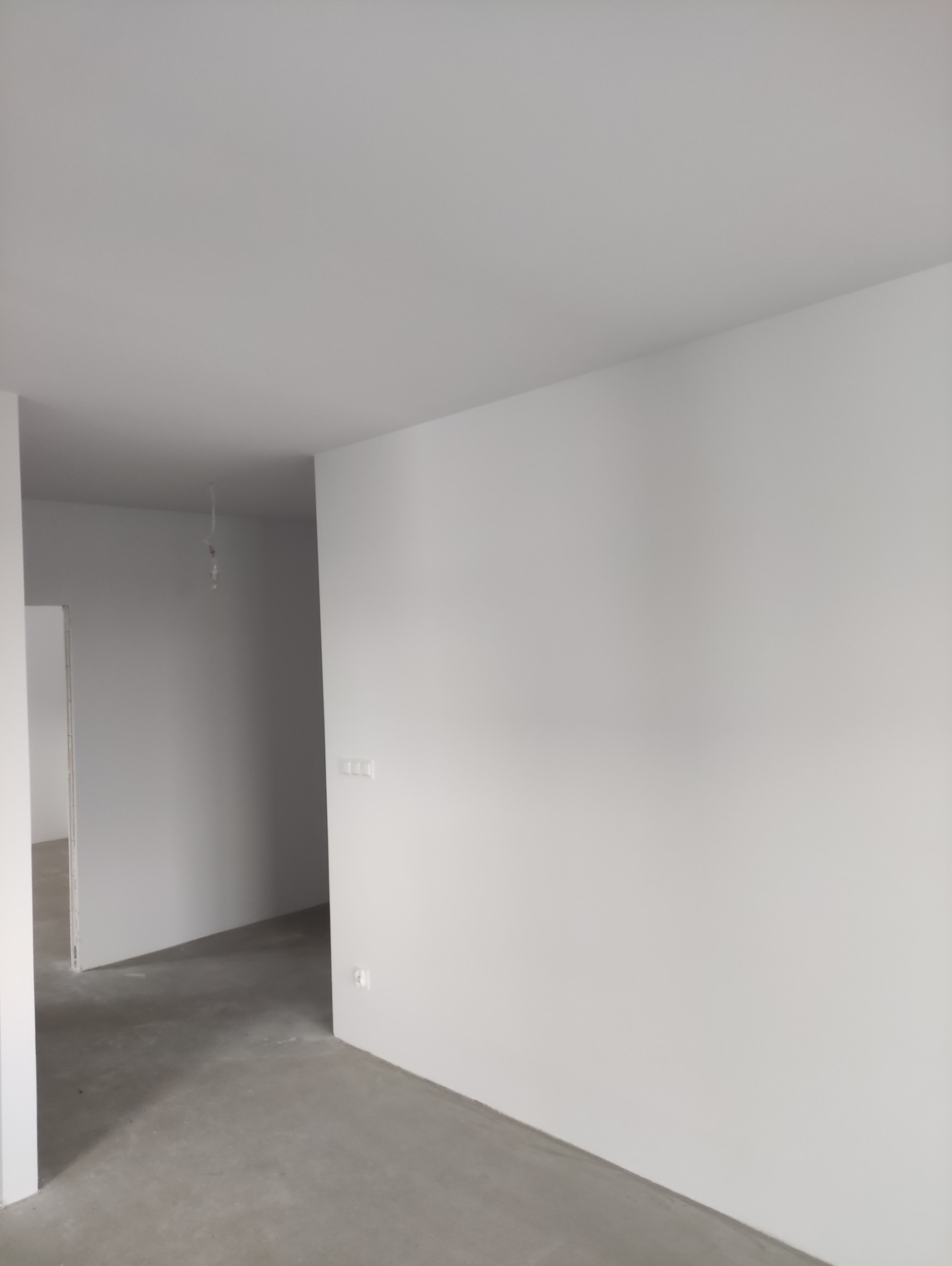 Wnętrze po remoncie: białe ściany, szara podłoga, widoczne gniazdka elektryczne. Minimalistyczny styl wykończenia, surowy beton na podłodze. W tle widoczne kolejne pomieszczenia.