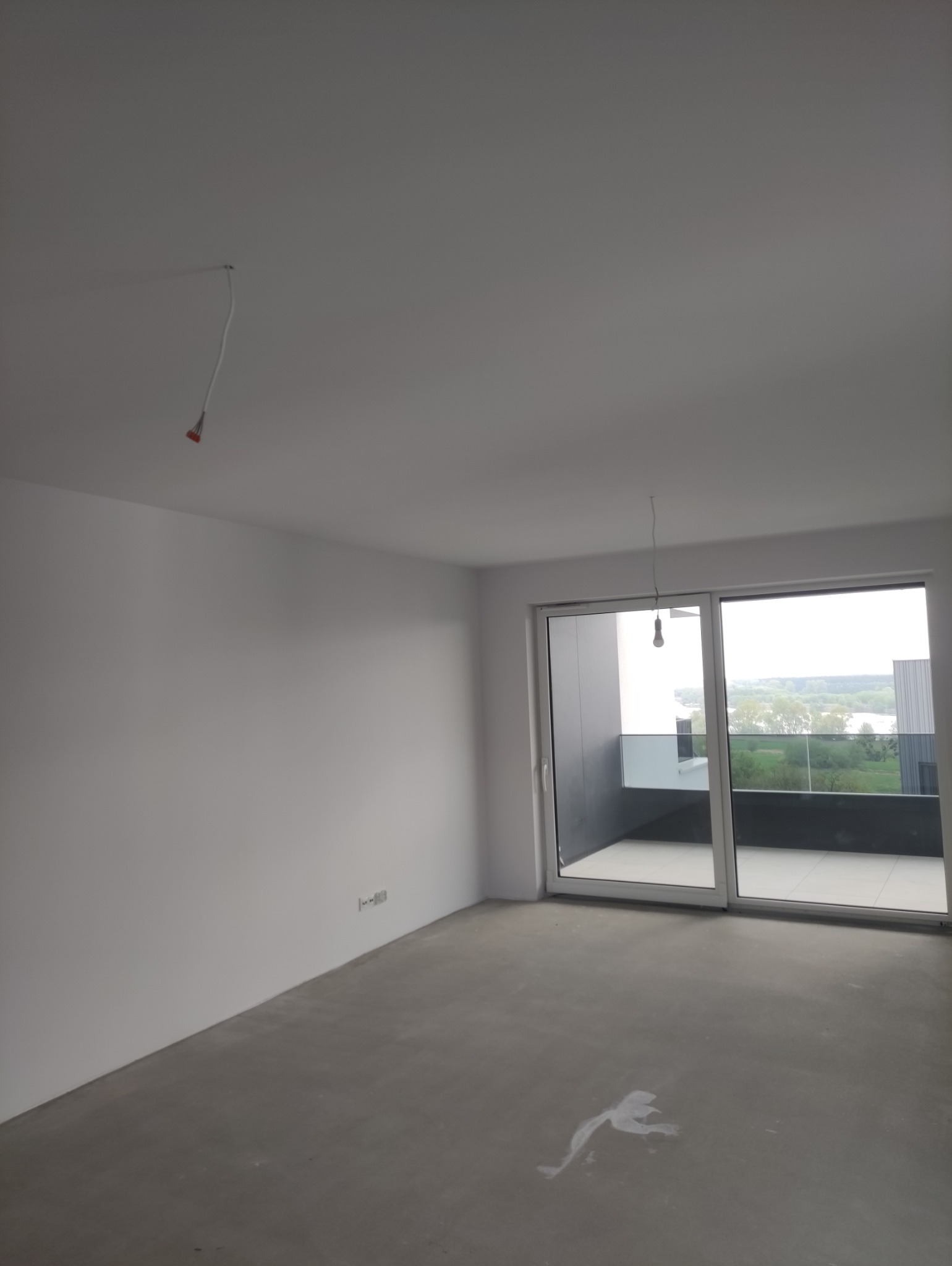 Wnętrze pokoju z gładkimi, białymi ścianami i surową podłogą. Widoczne gniazdka elektryczne, kabel zwisający z sufitu i okno balkonowe z widokiem na zieleń.