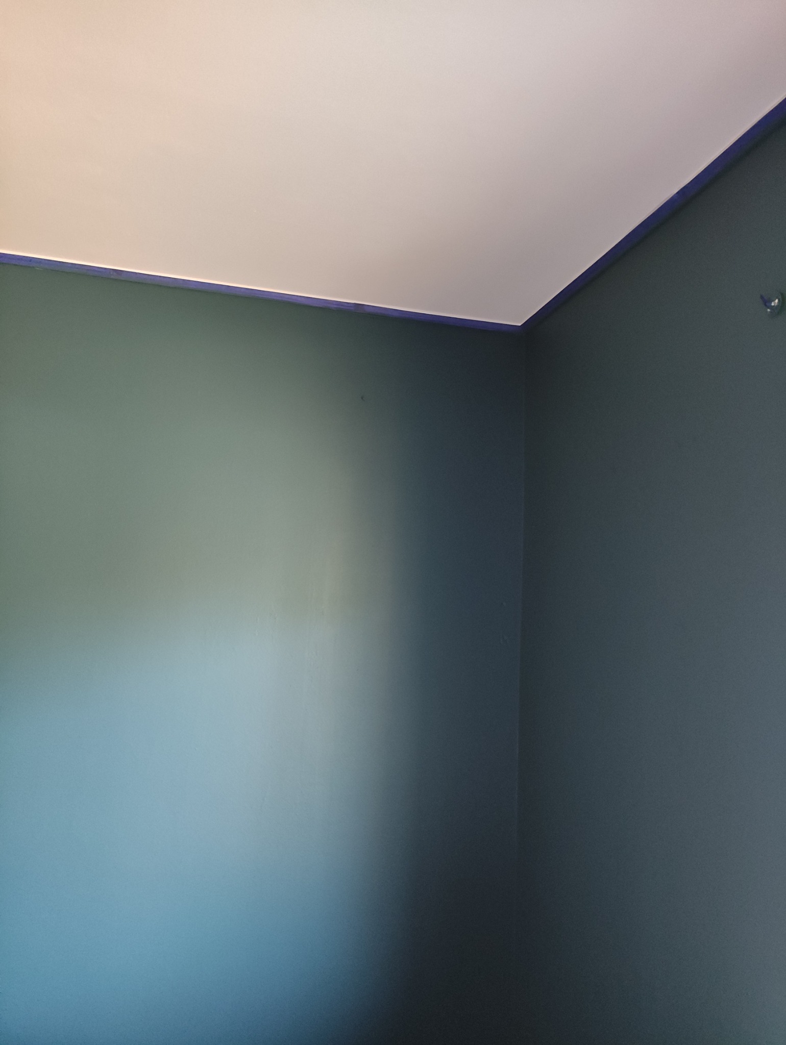 Pomieszczenie po malowaniu na szaro, z widocznym odcięciem od sufitu zabezpieczonym taśmą malarską. Świeżo odmalowane ściany, narożnik pokoju.