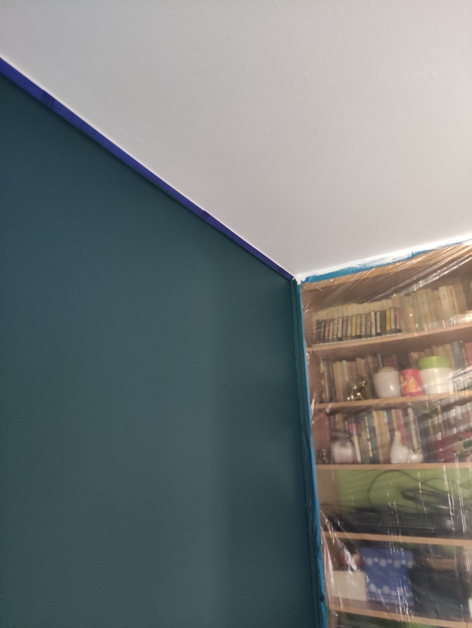 Ściana pomalowana na ciemny turkus, zabezpieczona taśmą malarską przy suficie. Regał z książkami osłonięty folią ochronną.