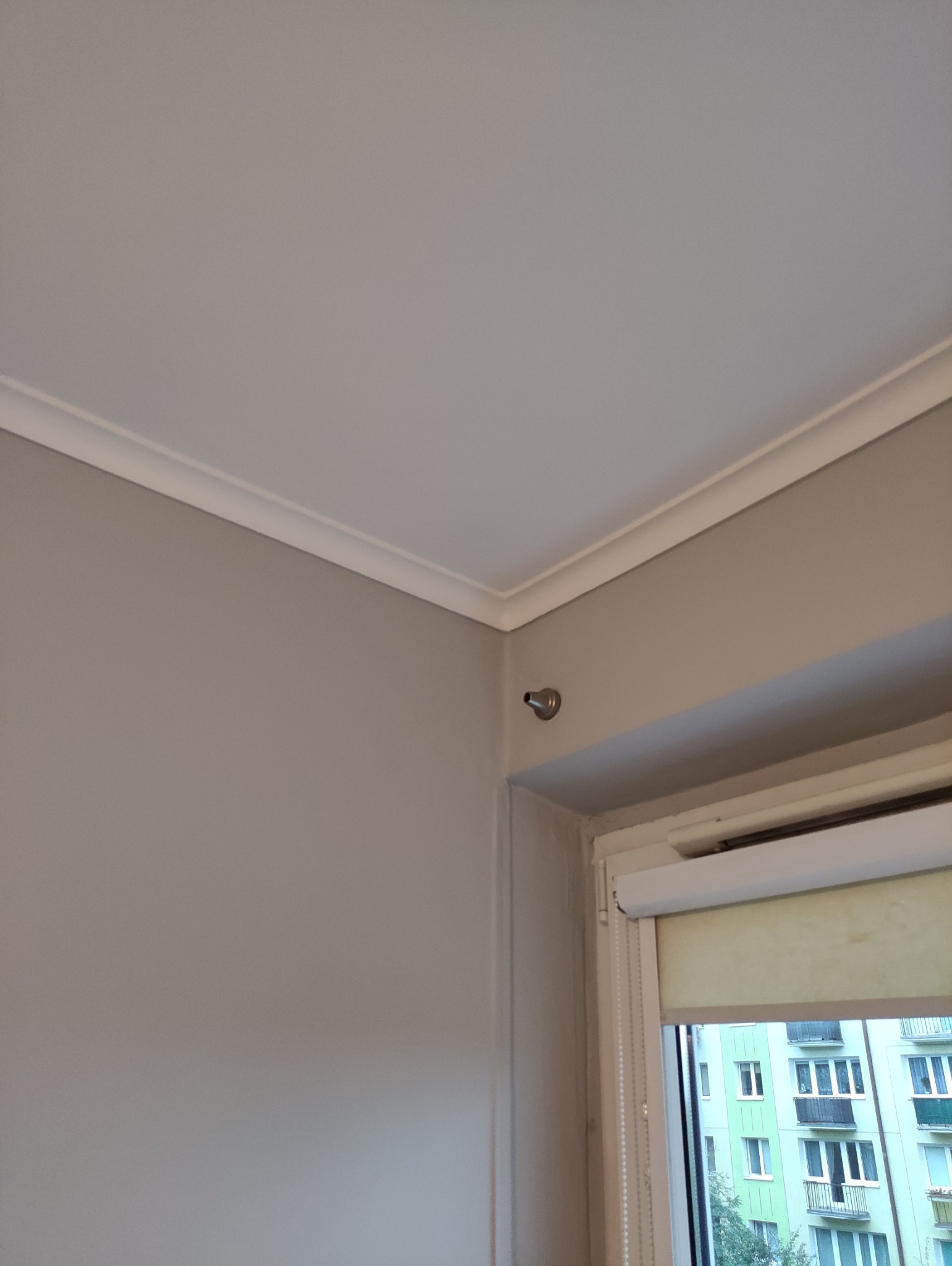 Pokój po odświeżeniu: ściany pomalowane na szaro, biały sufit z dekoracyjną listwą przy suficie, okno z roletą w tle. Widoczny fragment ściany i sufitu.
