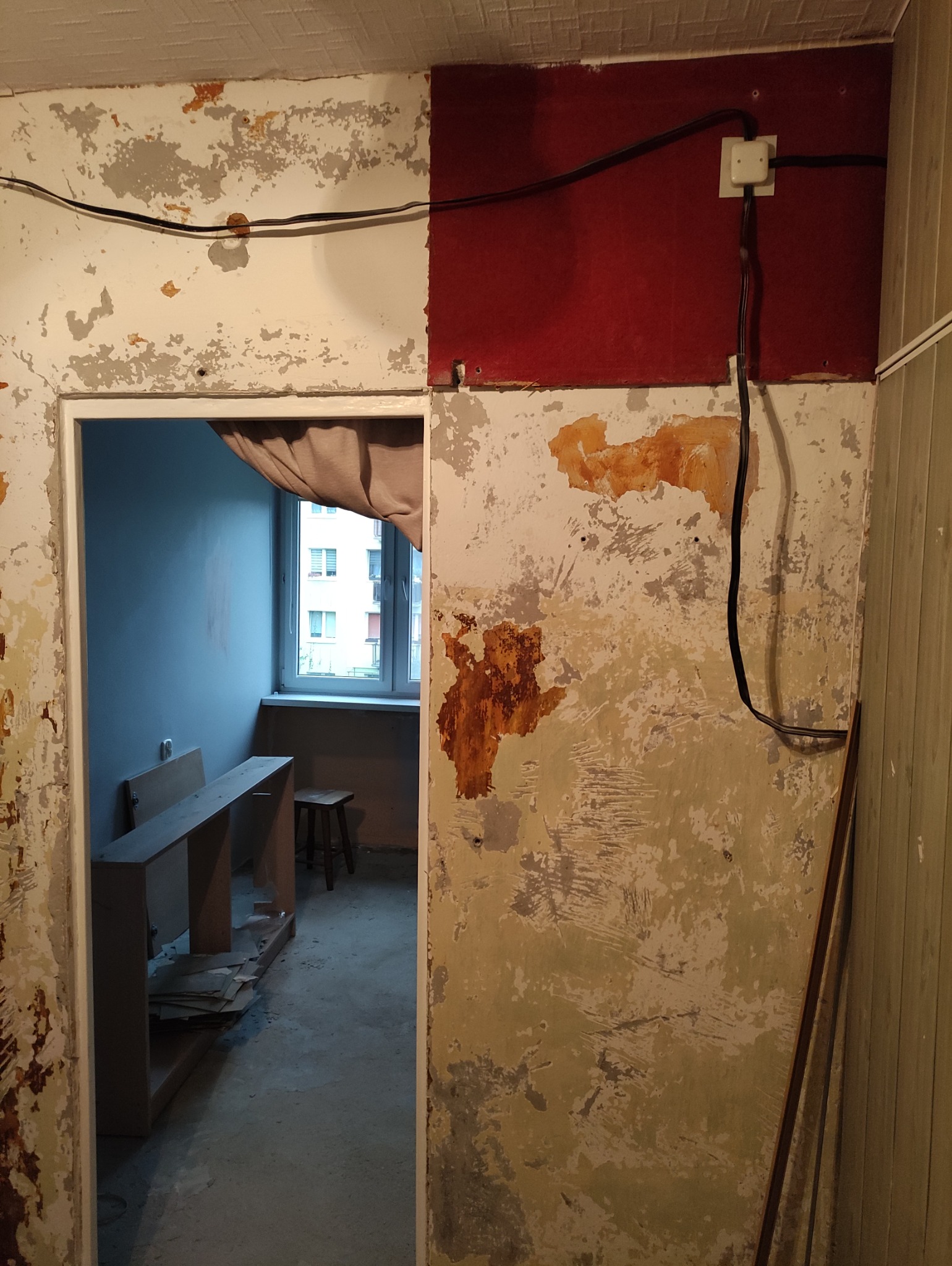 Pokój przed remontem: zniszczone ściany z resztkami farby, widoczne kable i instalacja elektryczna. W tle okno z widokiem na budynek.