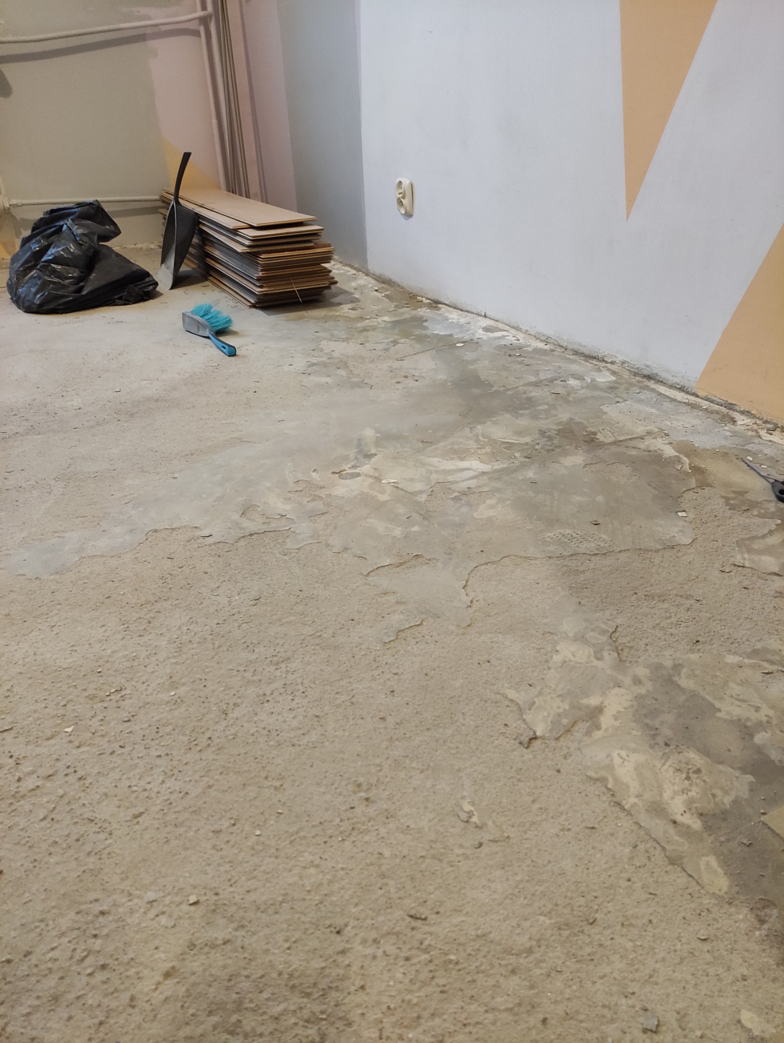 Przygotowanie podłoża pod panele: nierówna wylewka, deski paneli, zmiotka i worek na śmieci w rogu pokoju. Widoczne gniazdko i fragment ściany z malowaniem.