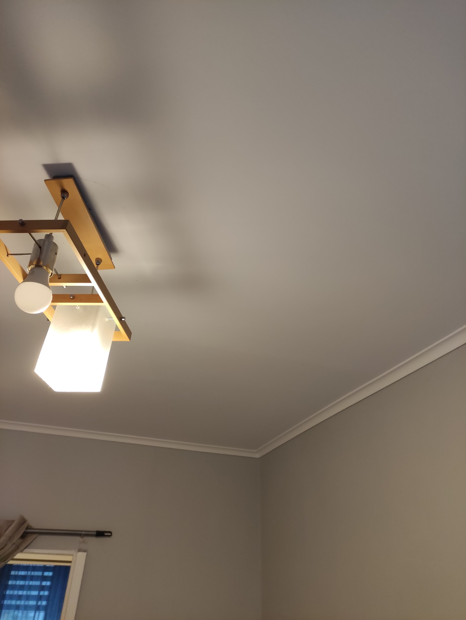 Szare wnętrze pokoju z drewnianą lampą sufitową i listwą dekoracyjną pod sufitem. Widoczny fragment okna z zasłoną.