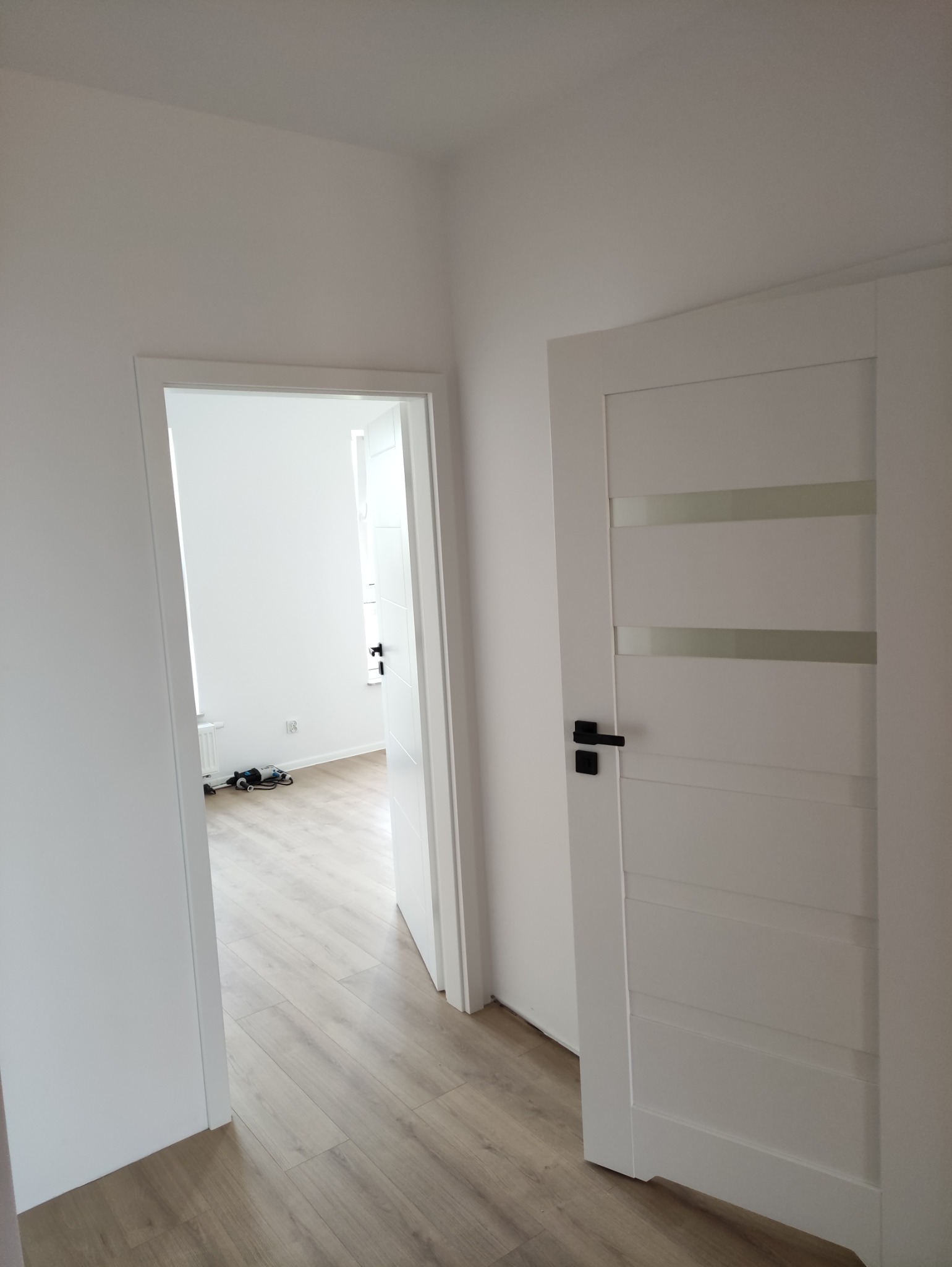 Nowoczesne, białe drzwi z czarną klamką i przeszklonymi wstawkami, widoczne na tle świeżo pomalowanego, jasnego wnętrza z panelami podłogowymi.