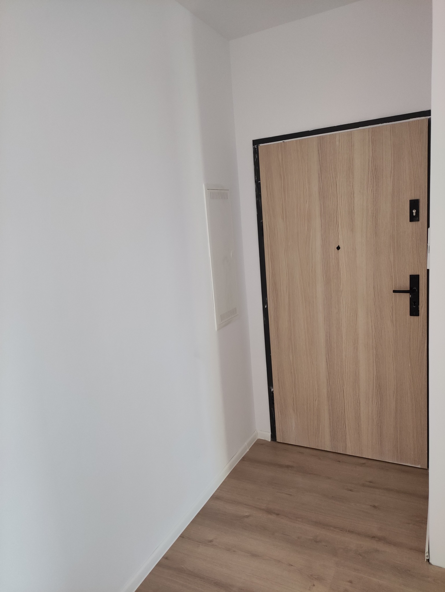 Jasne wnętrze po remoncie: białe ściany, panele podłogowe w odcieniach brązu i drzwi wejściowe z okleiną imitującą drewno. Minimalistyczny design.