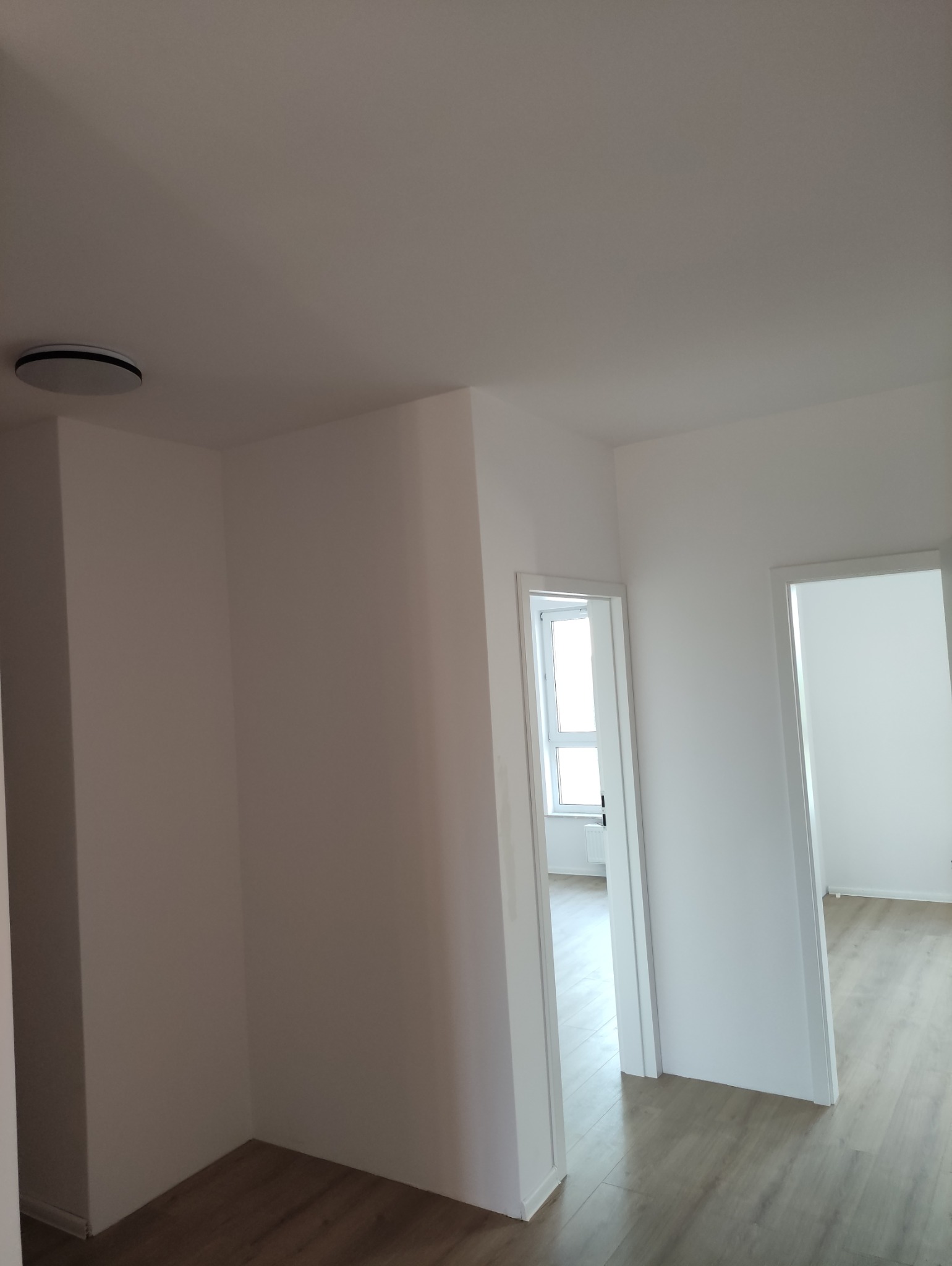 Białe, świeżo pomalowane wnętrze mieszkania z drewnopodobną podłogą, widok z korytarza na dwa pokoje z oknami. Minimalistyczny design, nowoczesne oświetlenie sufitowe.