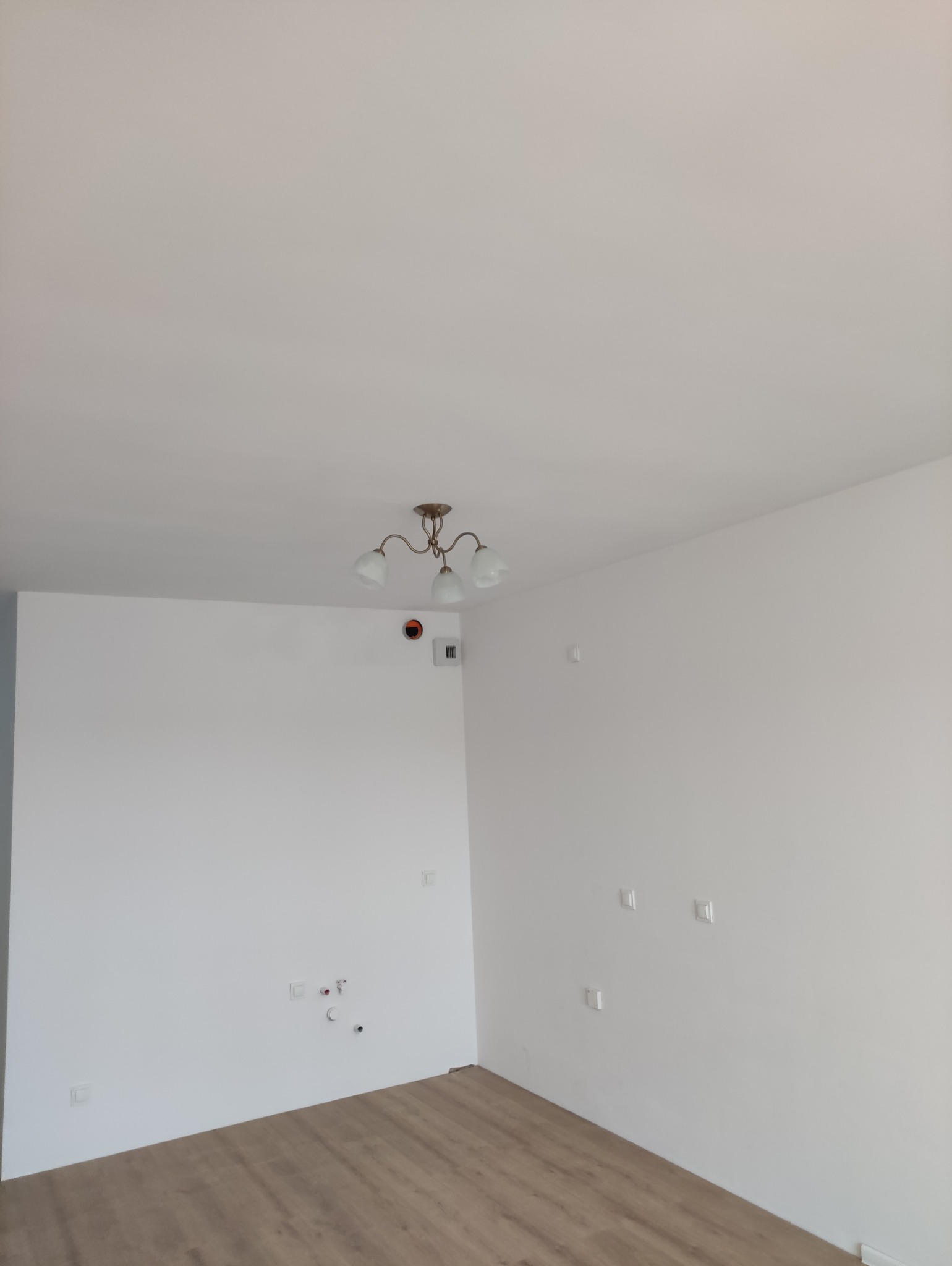 Jasne, odnowione wnętrze pokoju z panelami podłogowymi i białymi ścianami. Widoczna instalacja elektryczna i oświetlenie sufitowe.