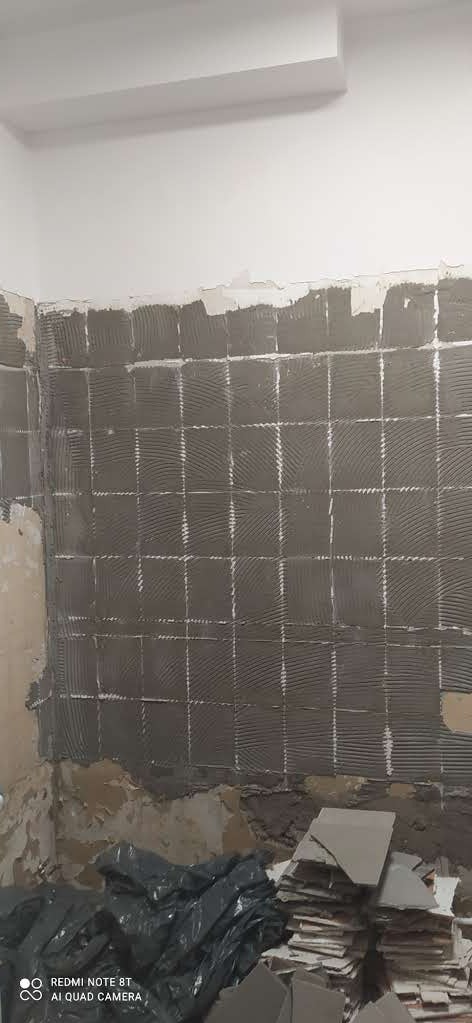 Ściana w trakcie układania płytek, widoczne klejone kwadraty i stos płytek na podłodze. Fragment ściany przygotowany pod płytki, sufit biały.