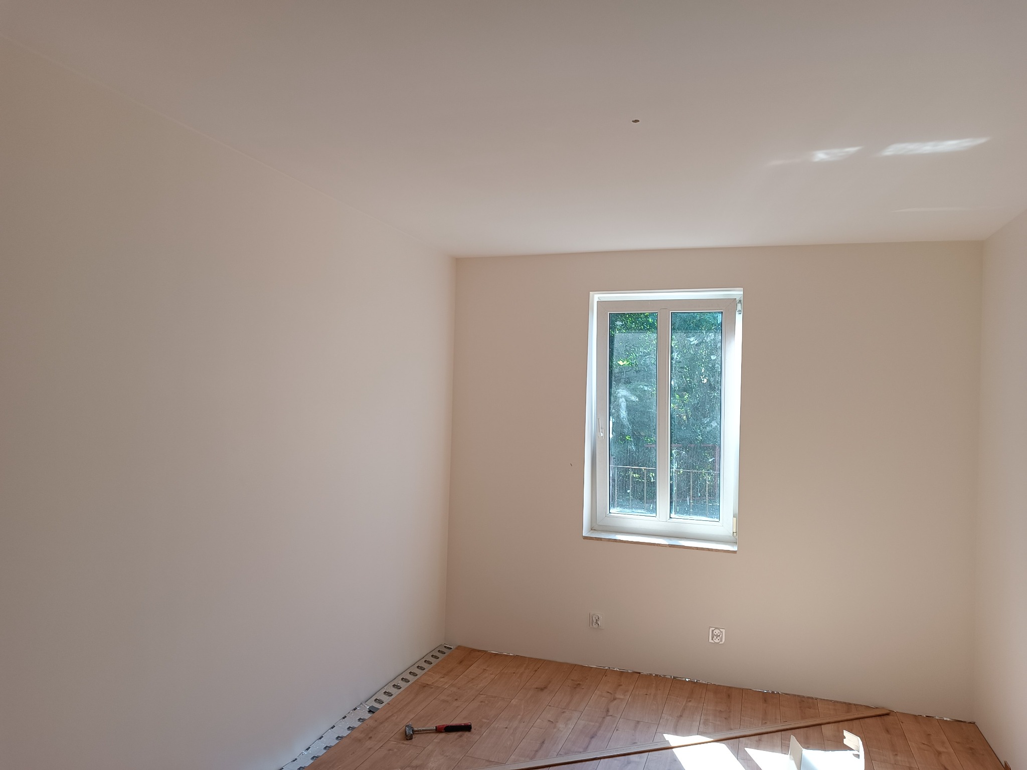 Jasne wnętrze po malowaniu z częściowo ułożoną podłogą z paneli; widoczne panele, listwy przypodłogowe, młotek i fragment okna.