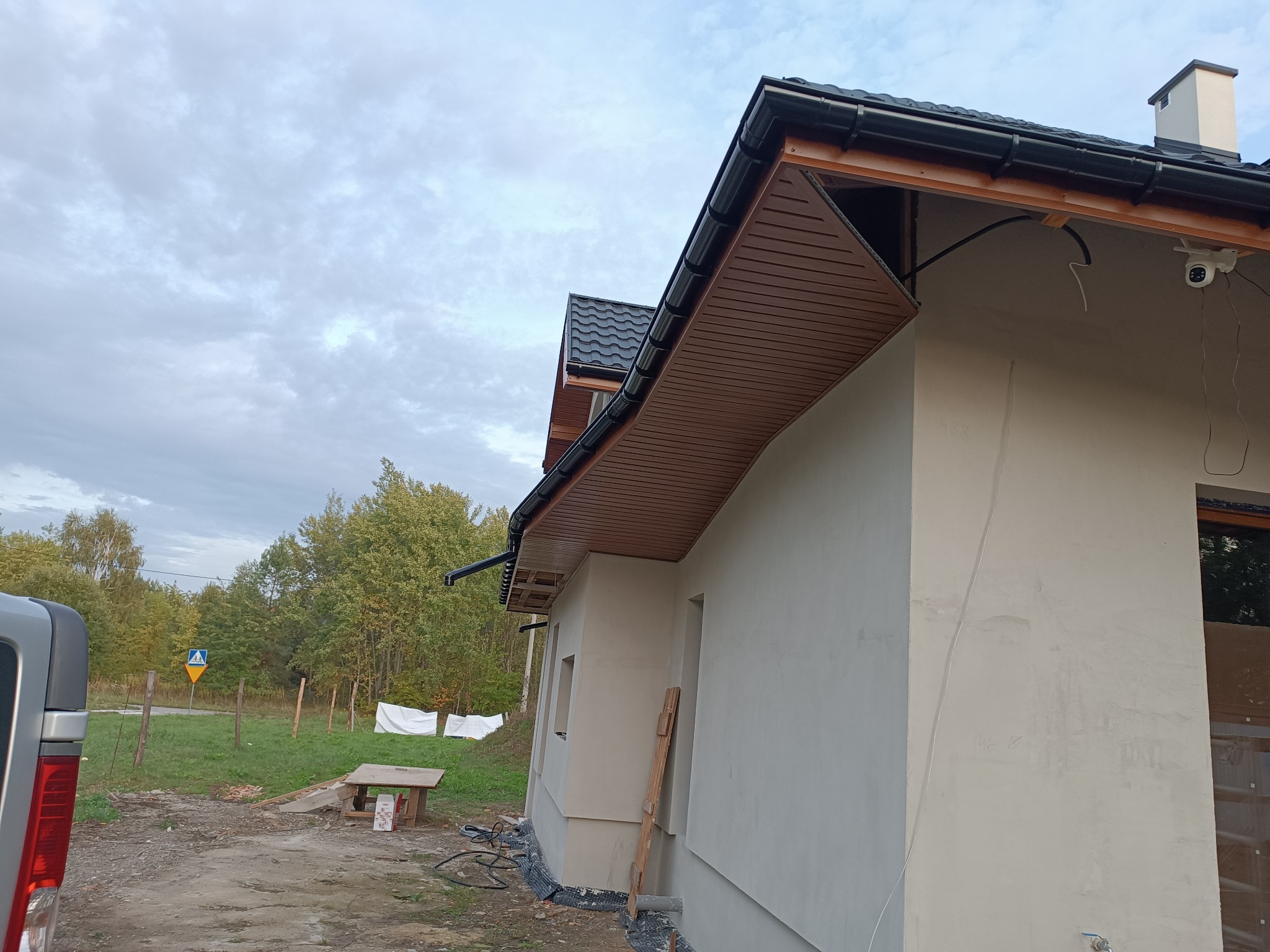 Fragment elewacji domu w trakcie wykańczania, widoczne orynnowanie, podbitka, instalacja kamery monitoringu oraz częściowo otynkowana ściana zewnętrzna.