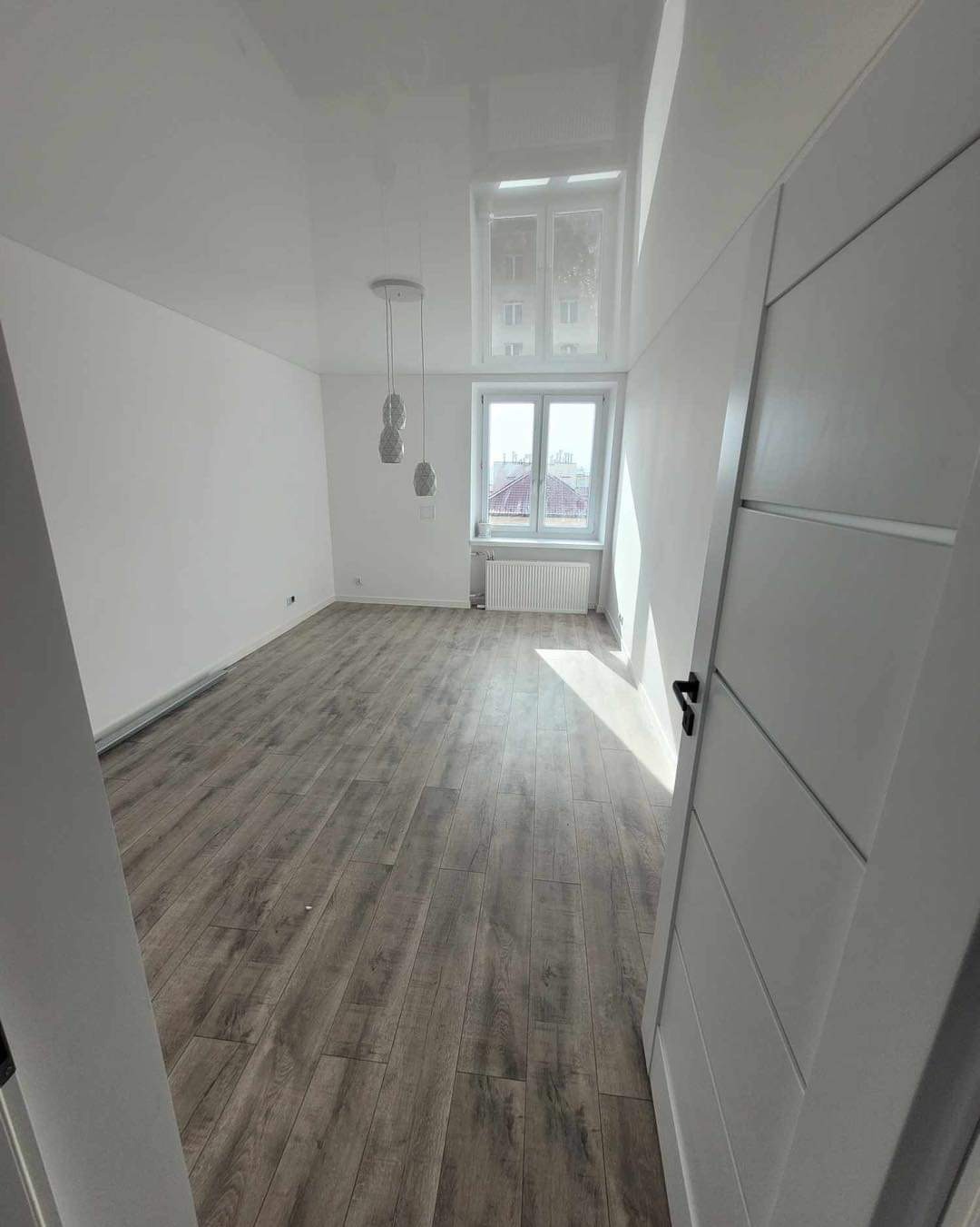 Jasne wnętrze z drewnopodobną podłogą z paneli, białymi ścianami, błyszczącym sufitem i nowoczesnym oświetleniem wiszącym. Częściowo widoczne drzwi z czarną klamką.