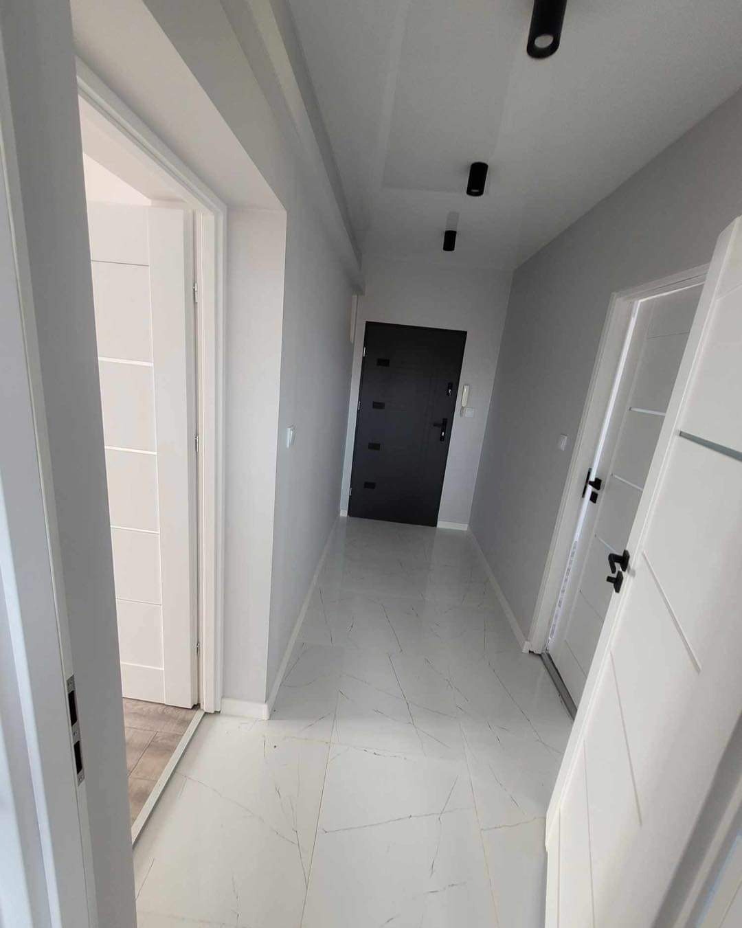 Wnętrze korytarza po remoncie: białe płytki podłogowe z szarymi żyłkami, szare ściany, czarne drzwi wejściowe, białe drzwi wewnętrzne z czarnymi klamkami, oświetlenie punktowe na suficie.