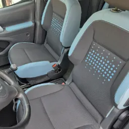 Wnętrze samochodu z widocznymi przednimi fotelami w kolorze szaro-błękitnym, z charakterystycznym wzorem z kwadratów na oparciach, widoczna kierownica i dźwignia zmiany biegów.
