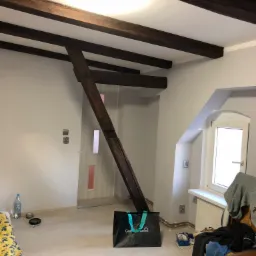 Pokój w Zielonej Górze z drewnianymi belkami stropowymi, częściowo pomalowanymi ścianami i elementami konstrukcyjnymi, widoczne niedociągnięcia i bałagan na podłodze.