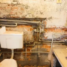 Odkryta instalacja wodna i fragment ściany z cegły w trakcie remontu łazienki, widoczne rury, zawory, fragment toalety i narzędzia.