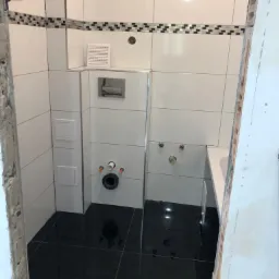 Wnętrze łazienki w trakcie prac: białe płytki na ścianach z dekoracyjnym pasem mozaiki, czarne płytki podłogowe, widoczne instalacje wodno-kanalizacyjne i fragment wanny, surowy otwór drzwiowy.