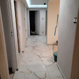 Korytarz w trakcie wykańczania: białe płytki imitujące marmur na podłodze, świeżo pomalowane ściany w odcieniach bieli i beżu, widoczne ślady po pracach wykończeniowych, taśma malarska na ścianie...