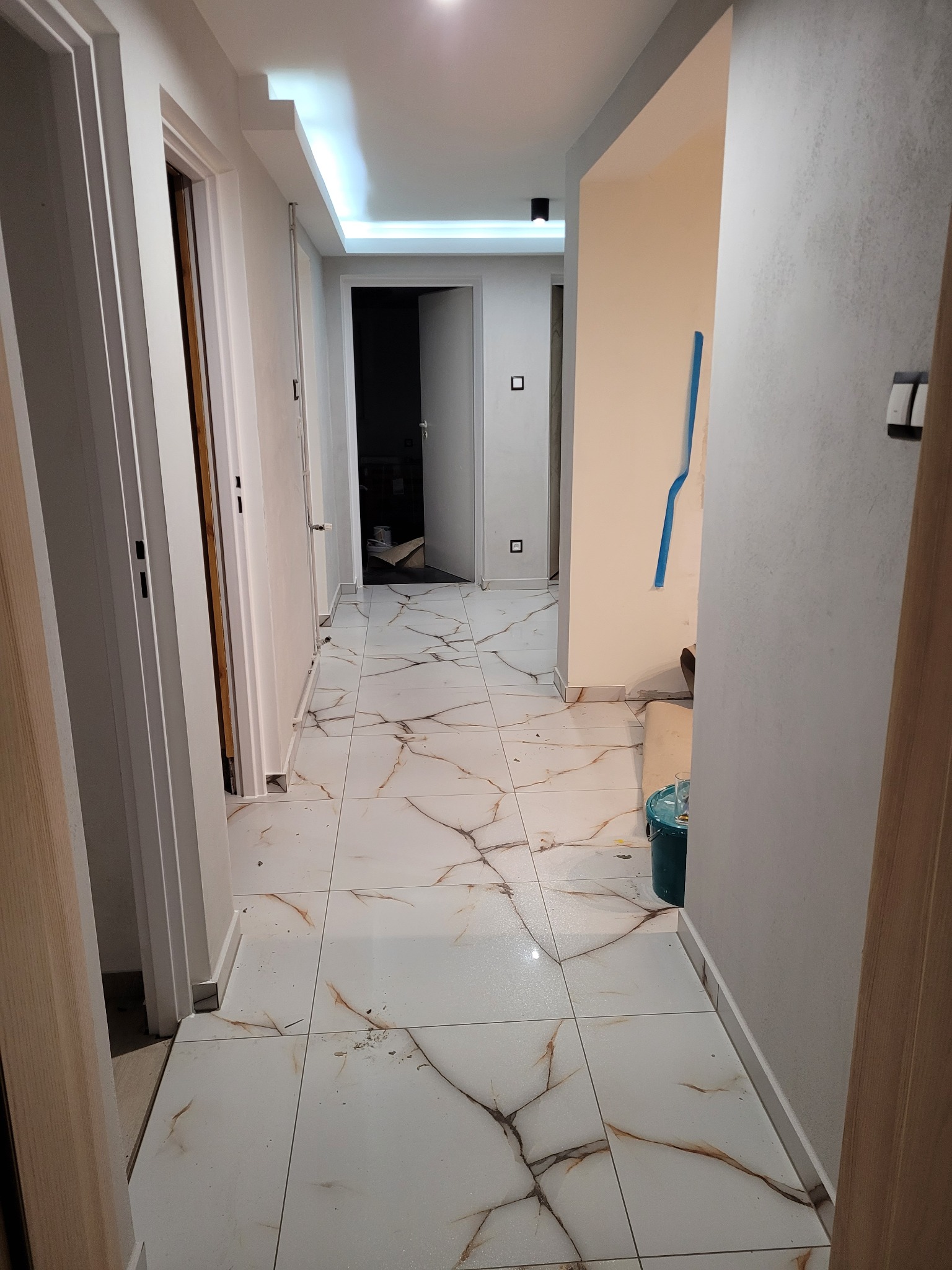 Korytarz w trakcie wykańczania: białe płytki imitujące marmur na podłodze, świeżo pomalowane ściany w odcieniach bieli i beżu, widoczne ślady po pracach wykończeniowych, taśma malarska na ścianie...
