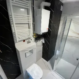 Nowoczesna łazienka po remoncie: białe płytki imitujące marmur, czarny akcent na ścianie, kabina prysznicowa i grzejnik drabinkowy, wisząca toaleta.