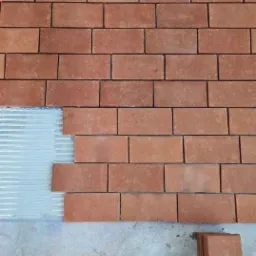 Ceglana ściana w trakcie układania, widoczny fragment z nałożonym klejem i stosem cegieł na podłodze.