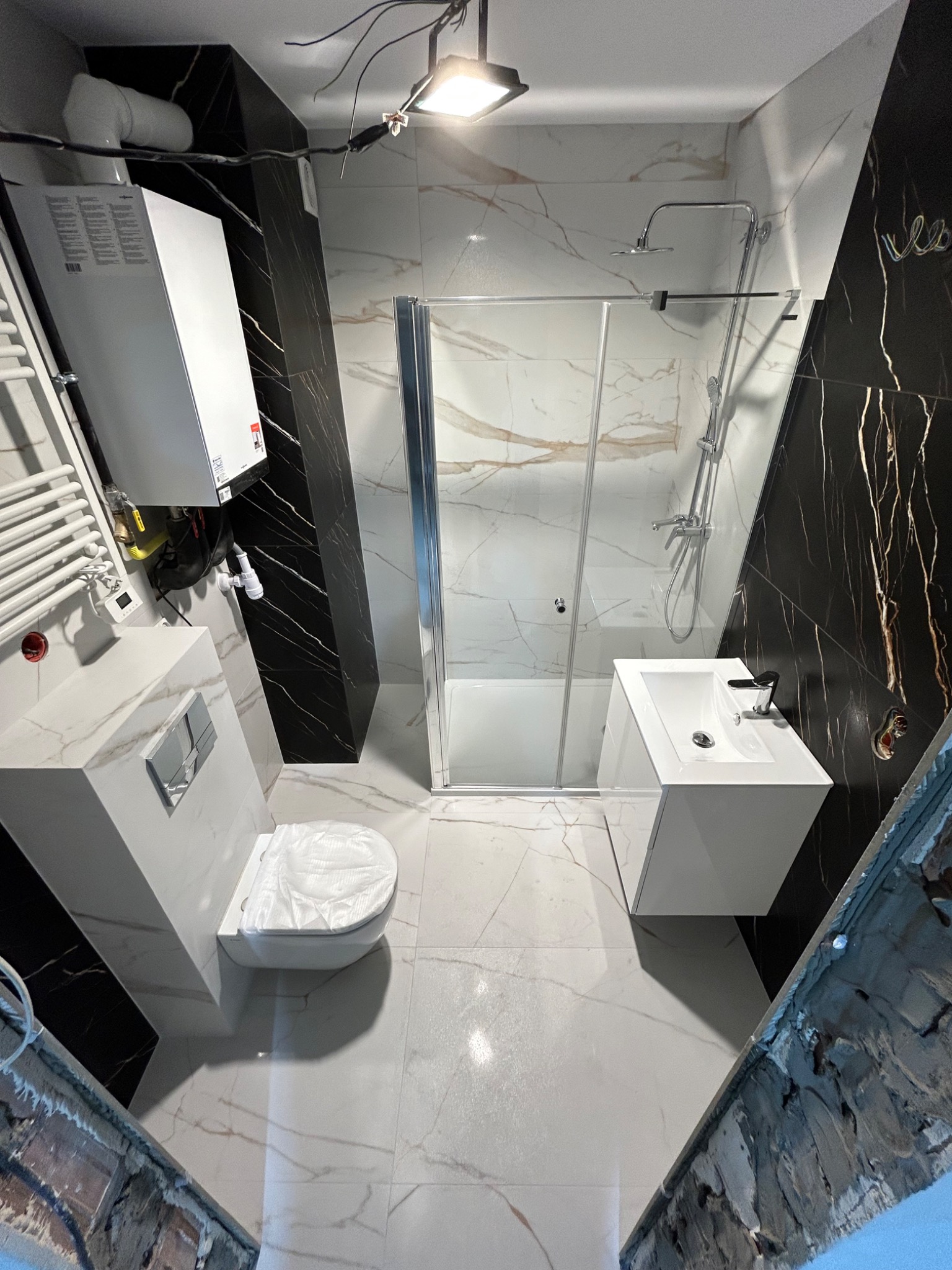 Wykończona łazienka z czarno-białymi płytkami imitującymi marmur, kabiną prysznicową, podwieszaną toaletą i bojlerem. Widoczne niedociągnięcia wykończeniowe.