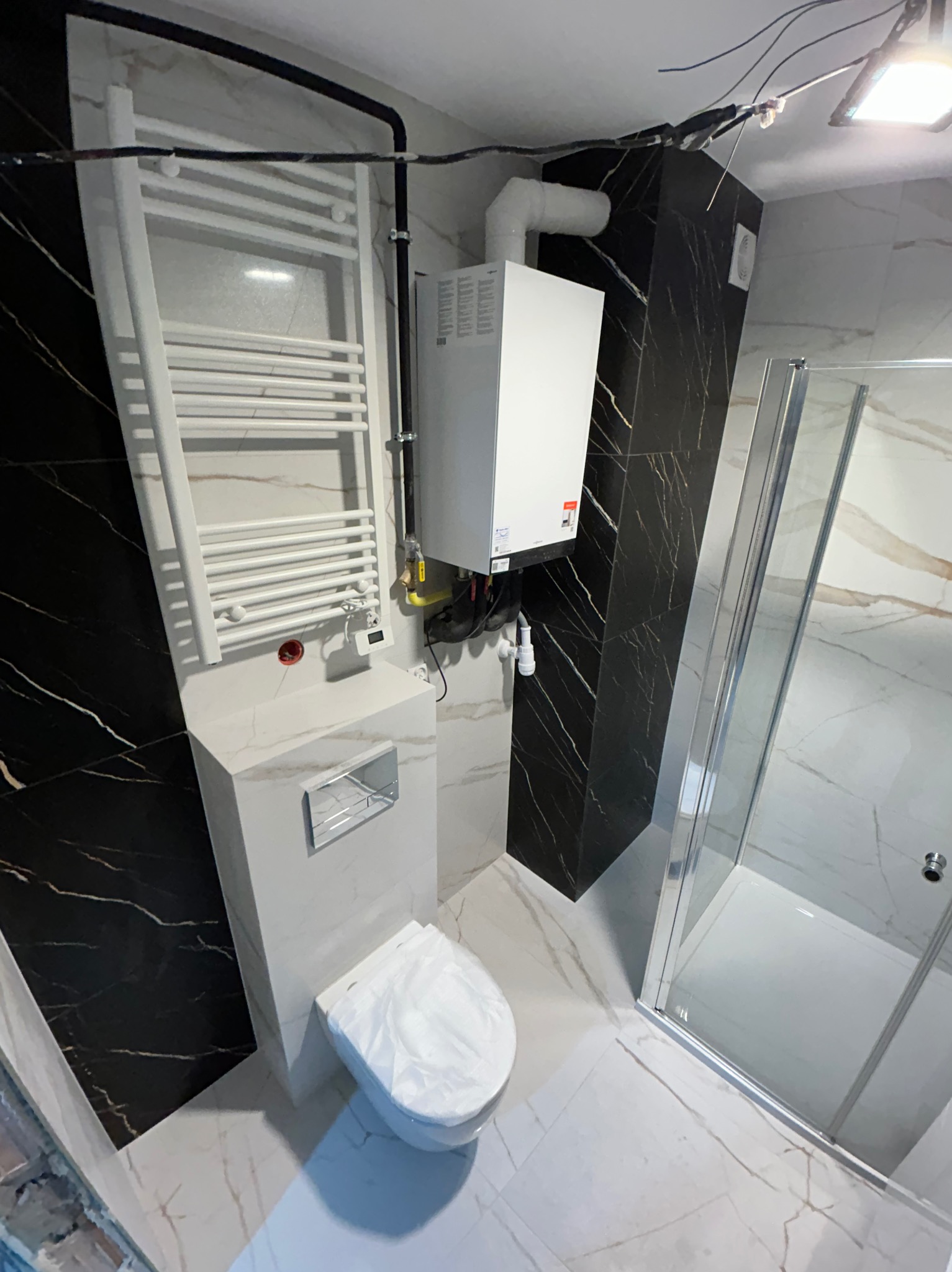 Nowoczesna łazienka po remoncie: białe płytki imitujące marmur, czarny akcent na ścianie, kabina prysznicowa i grzejnik drabinkowy, wisząca toaleta.