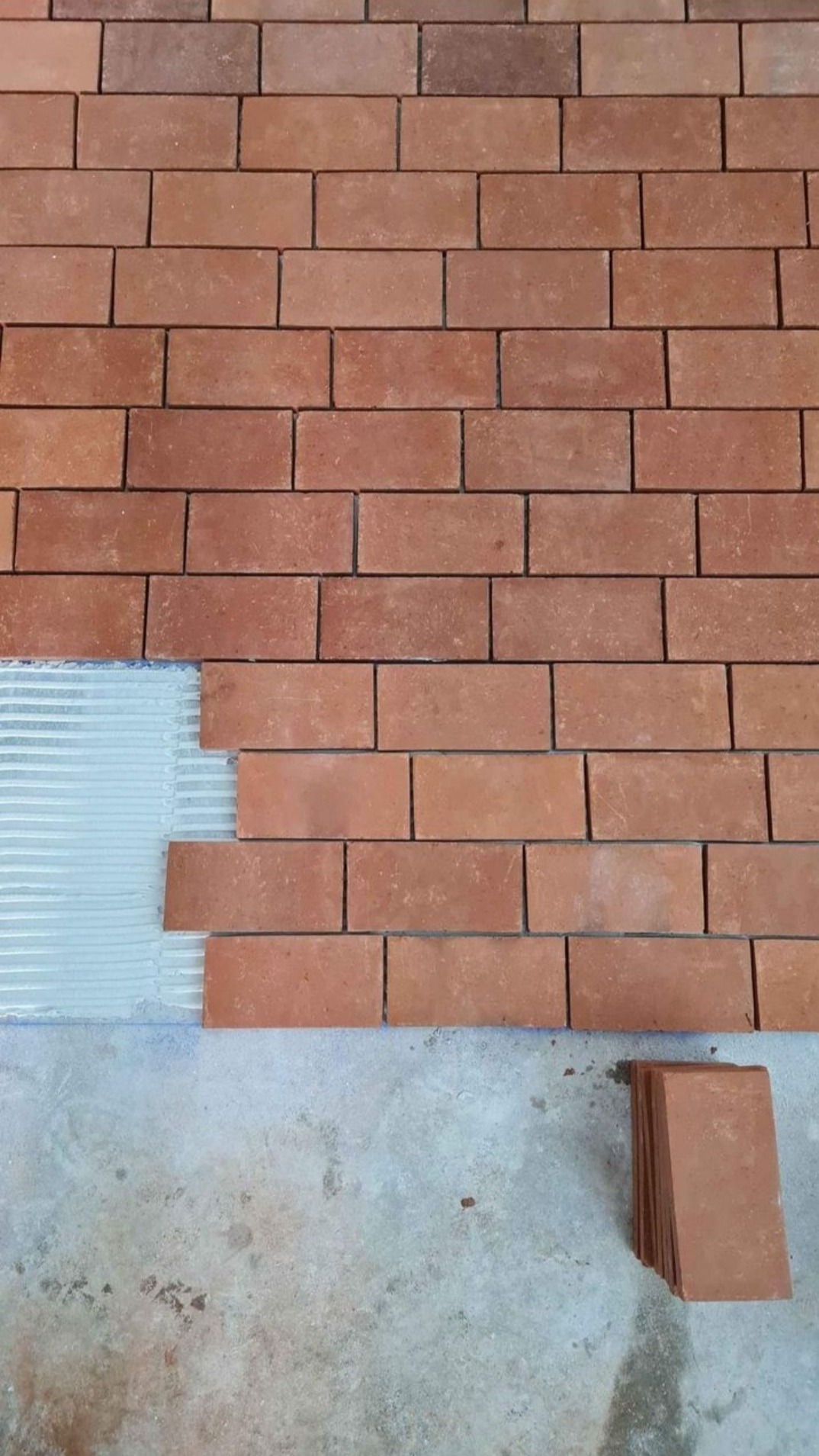 Ceglana ściana w trakcie układania, widoczny fragment z nałożonym klejem i stosem cegieł na podłodze.