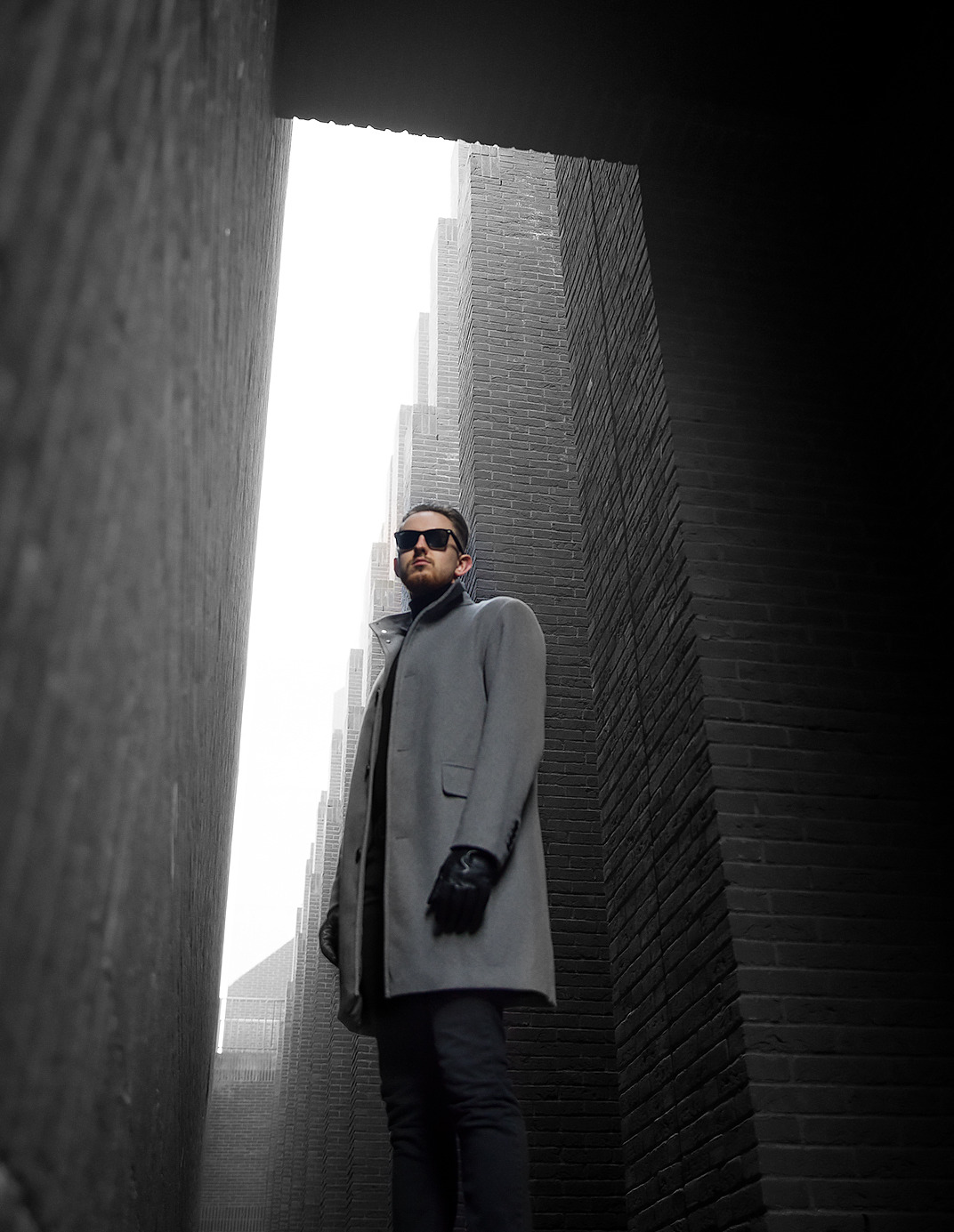 Mężczyzna w okularach przeciwsłonecznych i płaszczu, stojący w wąskiej przestrzeni między wysokimi budynkami z cegły, ujęcie od dołu.