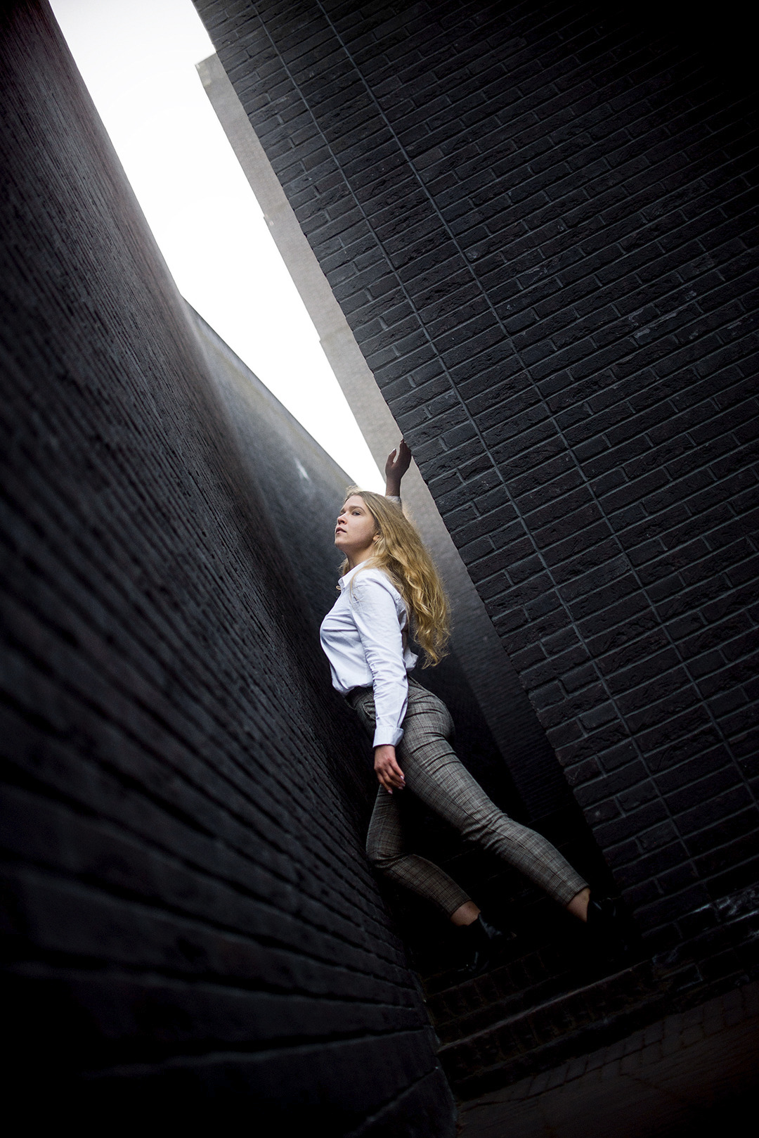 Młoda kobieta w białej koszuli i wzorzystych spodniach pozuje między wysokimi, ciemnymi ścianami z cegły w Gdańsku, spoglądając w górę w wąski pas światła.