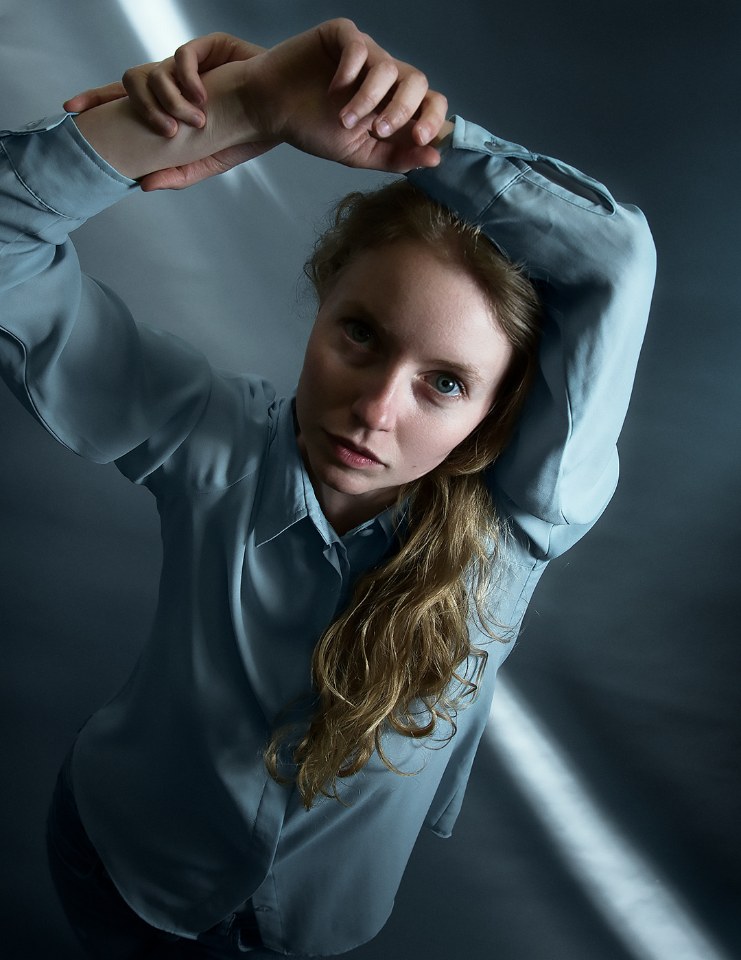 Portret kobiety w błękitnej koszuli, z rękami uniesionymi nad głową, patrzącej w dół z intensywnym spojrzeniem, oświetlonej przez pojedynczy strumień światła na tle szarego, gładkiego tła.