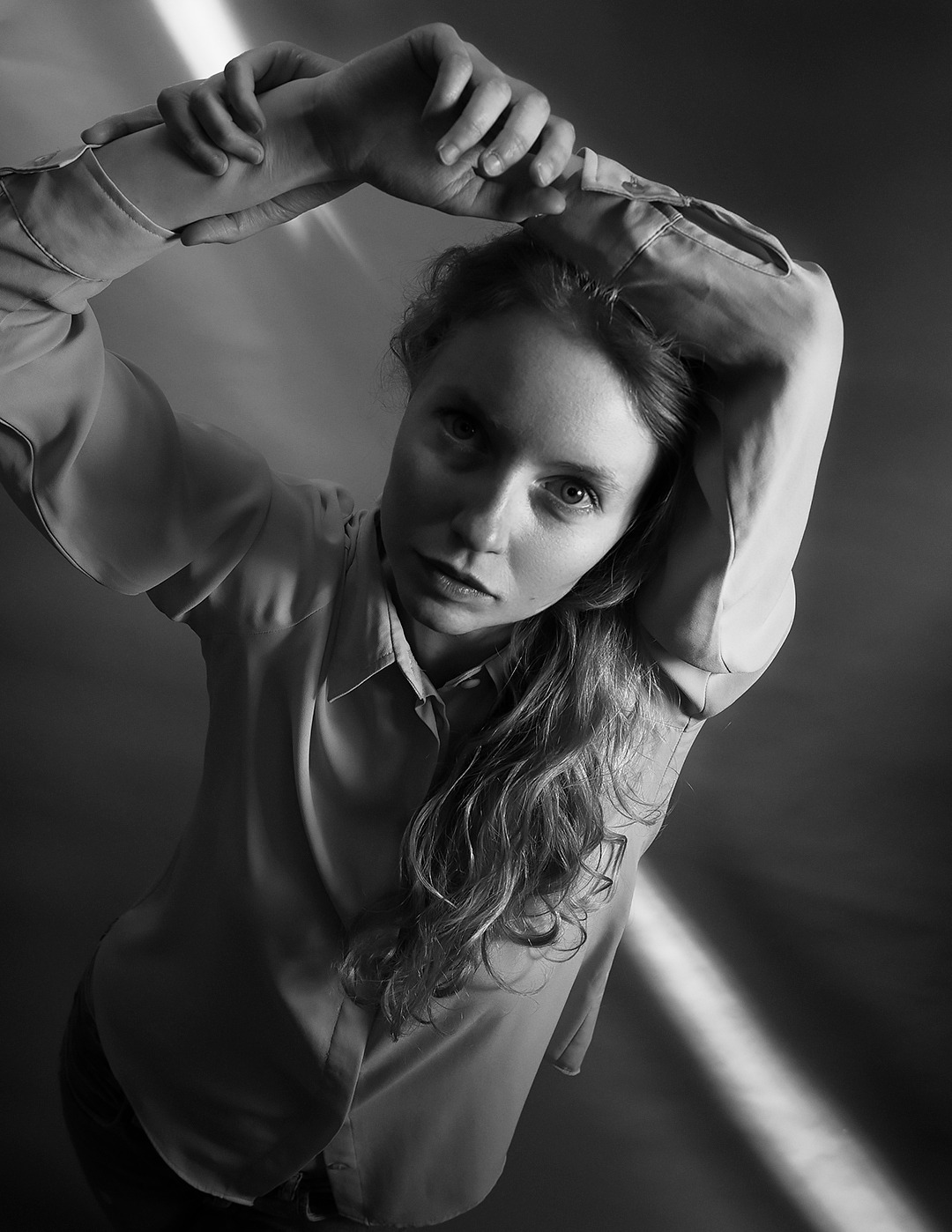 Portret kobiety w koszuli, z rękami uniesionymi nad głową, wykonany w czerni i bieli, z perspektywy żabiej, oświetlony pojedynczym promieniem światła.