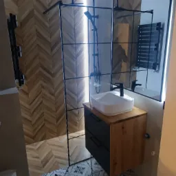 Nowoczesna łazienka z kabiną prysznicową typu walk-in z czarnymi profilami, umywalką nablatową na szafce z drewnianym blatem, wzorzystą podłogą i ścianą wyłożoną płytkami imitującymi drewno...