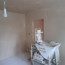 Wnętrze w trakcie prac, ściany pokryte świeżym tynkiem, widoczna maszyna tynkarska Bapro Strong Max 230V, surowa żarówka na kablu.