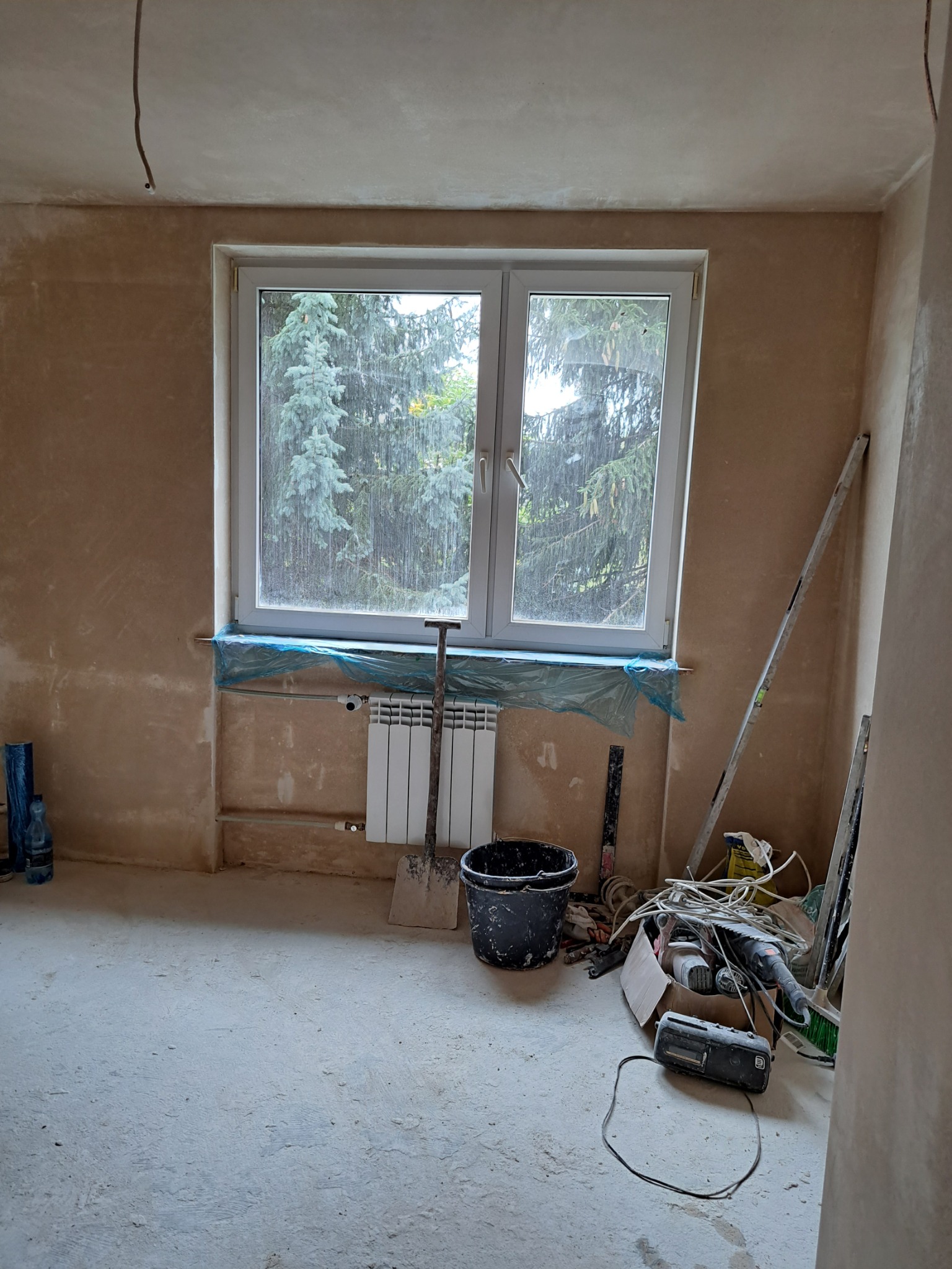 Pokój w trakcie prac wykończeniowych: ściany pokryte świeżym tynkiem, okno z widokiem na zieleń, zabezpieczony parapet folią, grzejnik, narzędzia i akcesoria budowlane na podłodze.