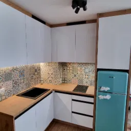 Nowoczesna kuchnia z białymi szafkami, drewnianymi blatami i kolorowymi płytkami nad blatem, turkusowa lodówka Amica z folią ochronną, drewnopodobna podłoga, czarna lampa sufitowa.