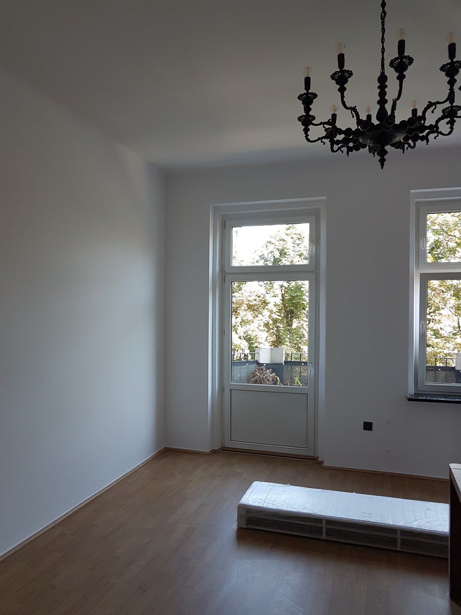 Pusty pokój z białymi ścianami, oknami balkonowymi, drewnianą podłogą i czarnym, ozdobnym żyrandolem, z zapakowanym w folię grzejnikiem na podłodze.