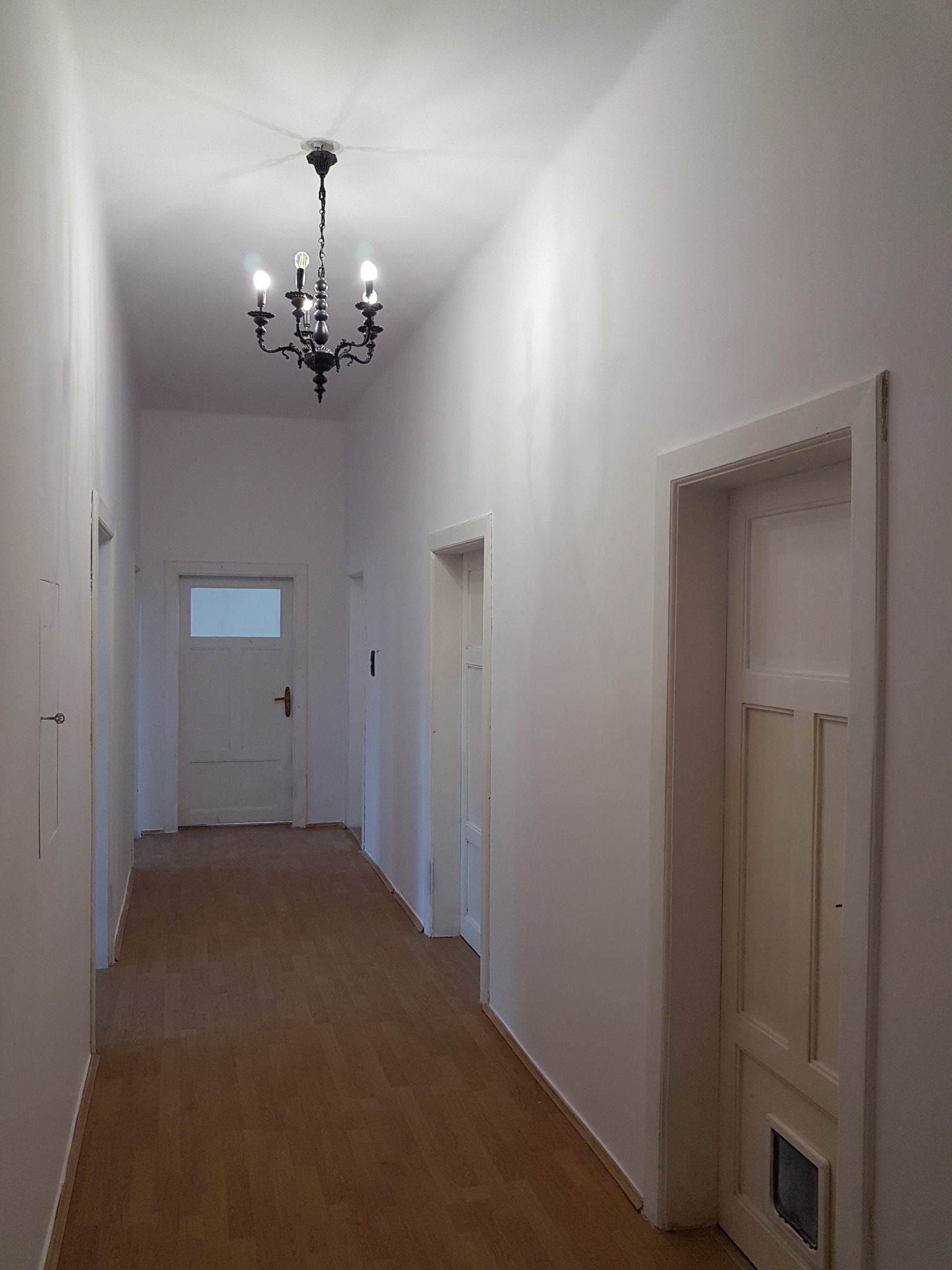 Długi, jasny korytarz po odświeżeniu, z drewnianą podłogą, białymi ścianami i drzwiami oraz czarnym, ozdobnym żyrandolem.